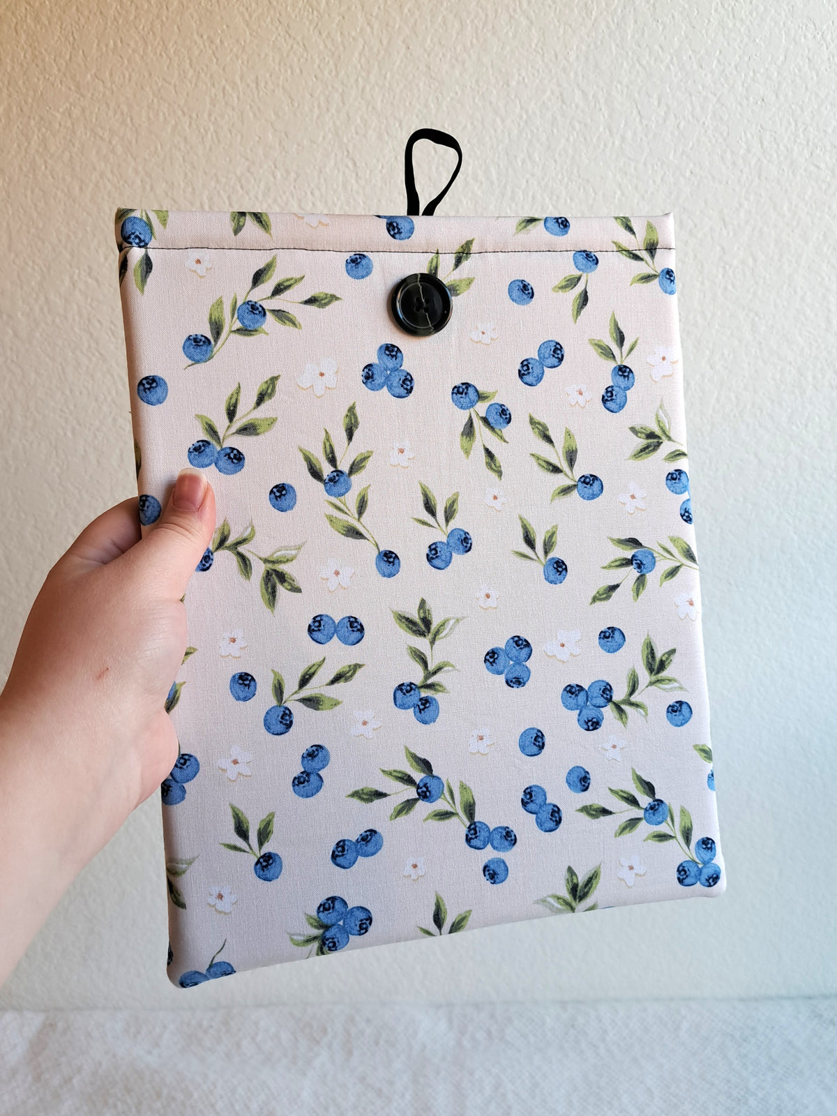 hand holding the booksleeve out. print is a light cream background with blueberries, leaves, and small white flowers spread out around the sleeve. Green button on front and black elastic on back