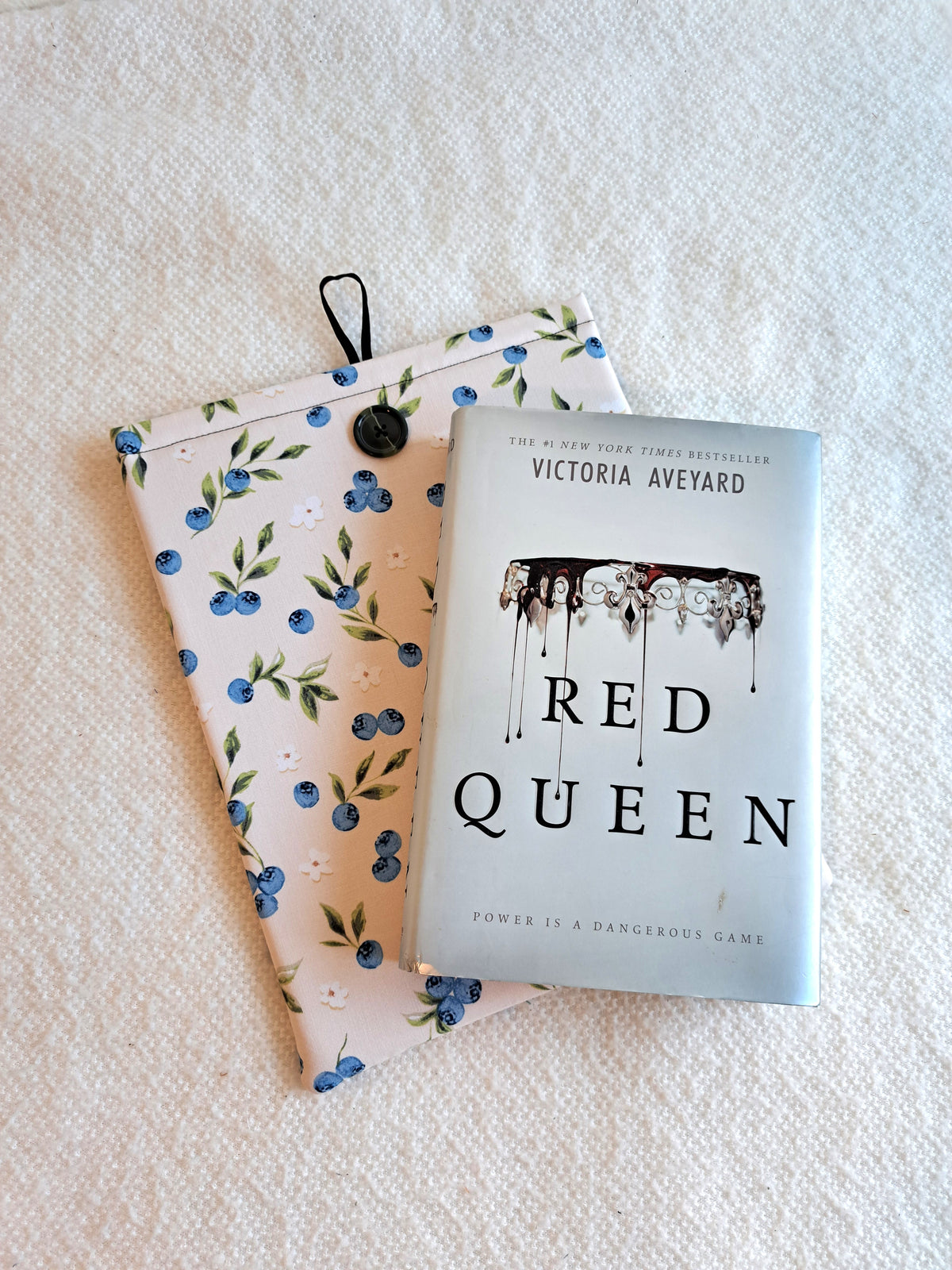 booksleeve is layed out on a white blanket the print is a light cream background with blueberries, leaves, and small white flowers spread out around the sleeve. Black button on front and black elastic on back. The book Red Queen is layed on top of the sleeve