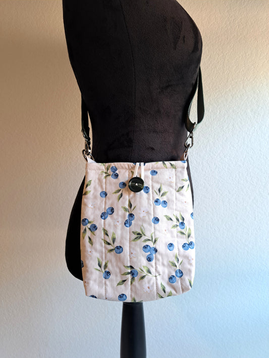 Bag is hanging on a black dressform with a white background. the bag print is a light cream background with blueberries, leaves, and small white flowers spread out around the sleeve, it is quilted with vertical lines. Green button on front and white elastic on back. Sage green strap with silver hardware.