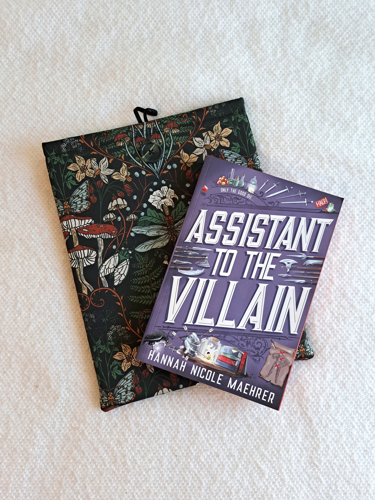 booksleeve is layed on a white blanket the print is on a dark background, a dragonfly motiff in the center surrounded by various mushrooms and plants in a dark earthy color palette. Black button on front and black elastic on back. The book Assistant to the Villian is layed on top of the booksleeve