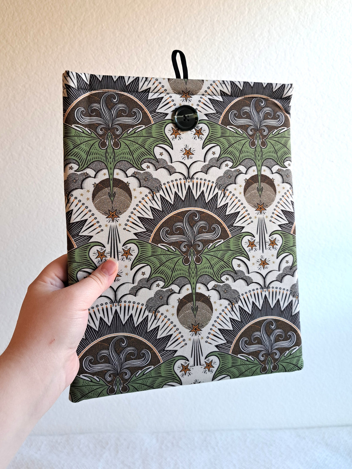 hand holding the booksleeve out. print is a repeated motiff of a green dragon with its wings spread out with a geometric sun in dark colors over it, illustrative clouds under it. and various stars and lines around it giving the look of the dragon flying. dark Green marbled button on front and black elastic on back
