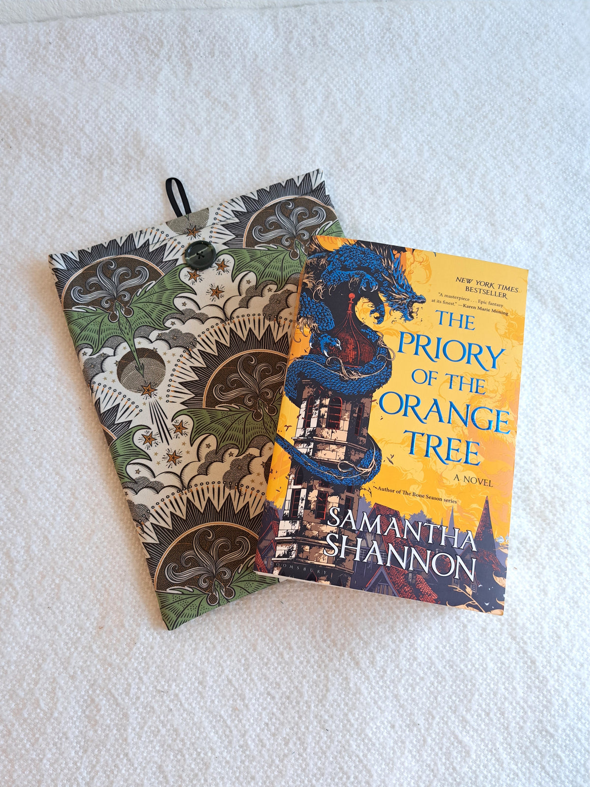 booksleeve is layed on a white blanket the print is a repeated motiff of a green dragon with its wings spread out with a geometric sun in dark colors over it, illustrative clouds under it. and various stars and lines around it giving the look of the dragon flying. dark Green marbled button on front and black elastic on back
the book Priory of the Orange Tree is layed on top of the sleeve.