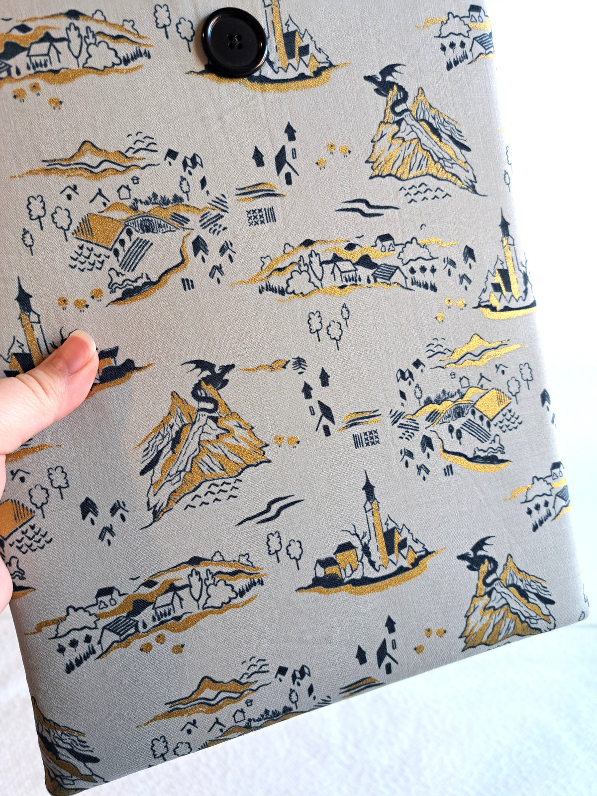 close up of hand holding the booksleeve out. print is a light gray background with black and gold metallic illustrative graphics. including a small dragon on mountains, a little town, part of a castle, tree, and mountains. Black button on front