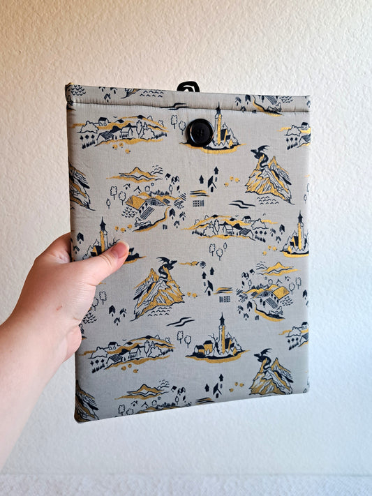 hand holding the booksleeve out. print is a light gray background with black and gold metallic illustrative graphics. including a small dragon on mountains, a little town, part of a castle, tree, and mountains. Black button on front and black elastic on back