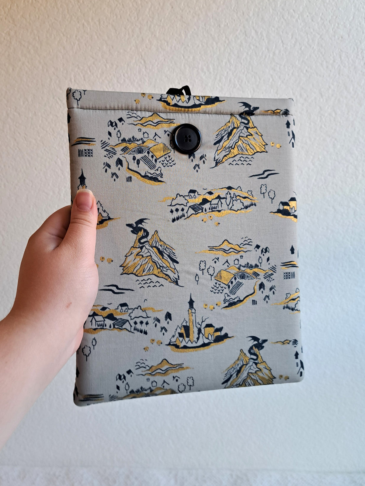 hand holding the booksleeve out. print is a light gray background with black and gold metallic illustrative graphics. including a small dragon on mountains, a little town, part of a castle, tree, and mountains. Black button on front and black elastic on back
