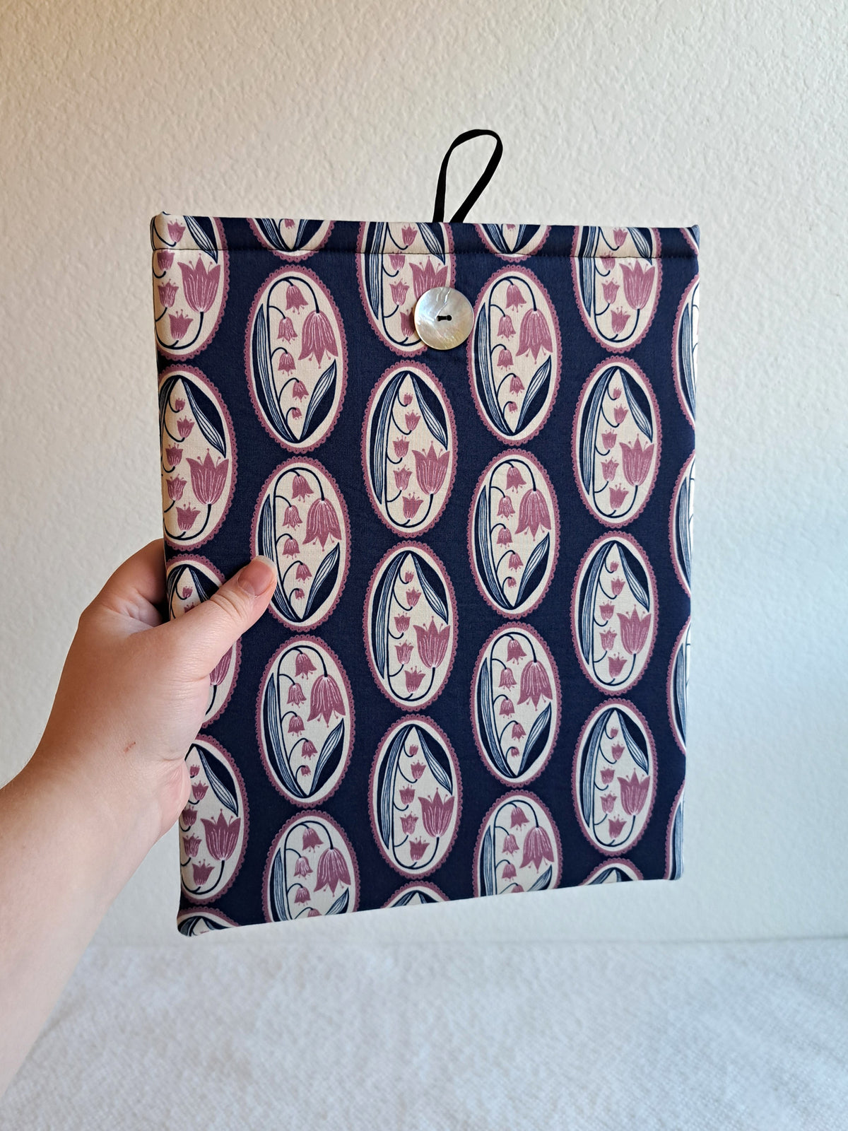 hand holding the booksleeve out. print is a repitition of a lilly of the valley flower in navy and a light purply pink inside of a white oval with a scallop boarder repeated in column on a navy blue background. Akoya shell button on front and black elastic on back