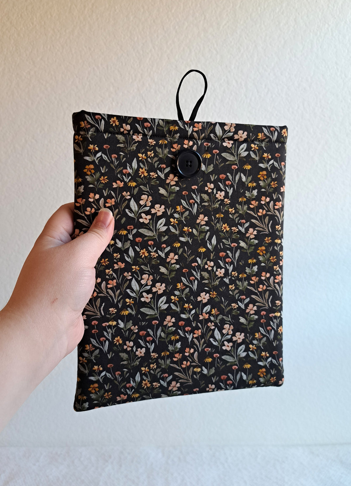 small booksleeve held out by a hand. The print has a dark background with dense small flowers and leaves in a non structured pattern. The flower colors consist of sage, light pink, yellow, terracotta, and red. Black button on front with black elastic on the back