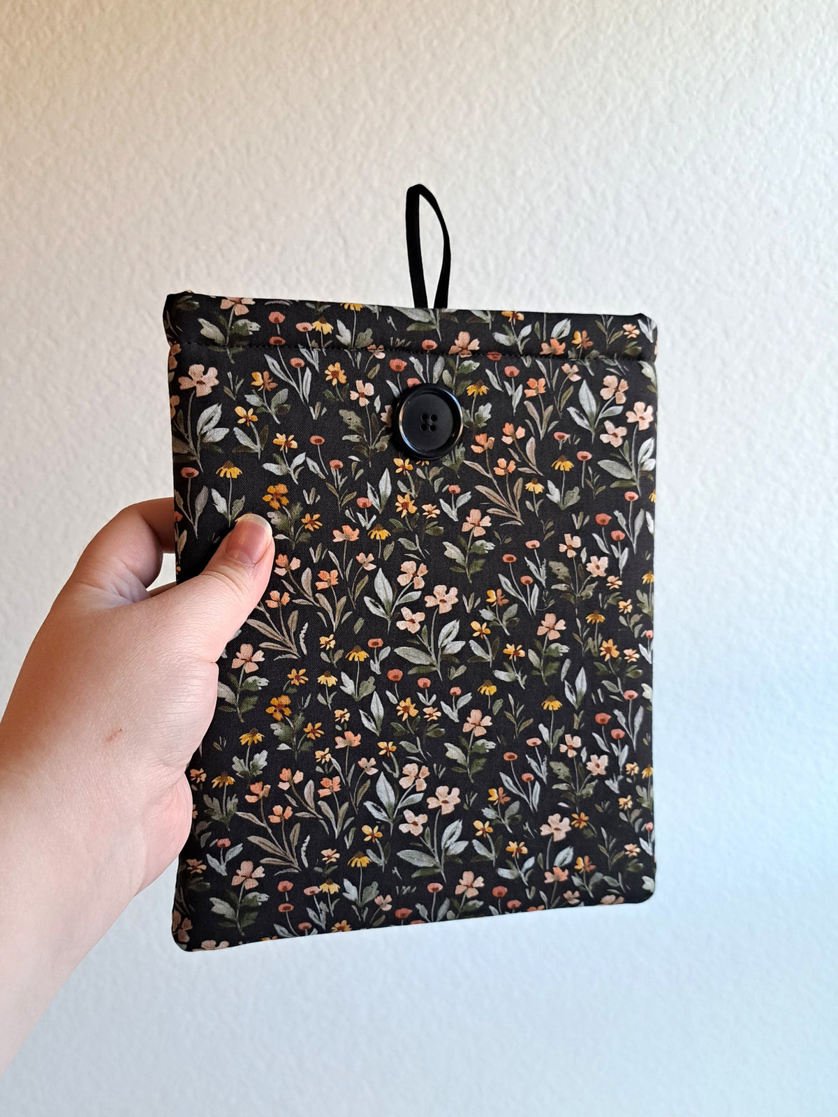 kinlde sleeve is held out by a hand. The print has a dark background with dense small flowers and leaves in a non structured pattern. The flower colors consist of sage, light pink, yellow, terracotta, and red. Black button on front with black elastic on the back