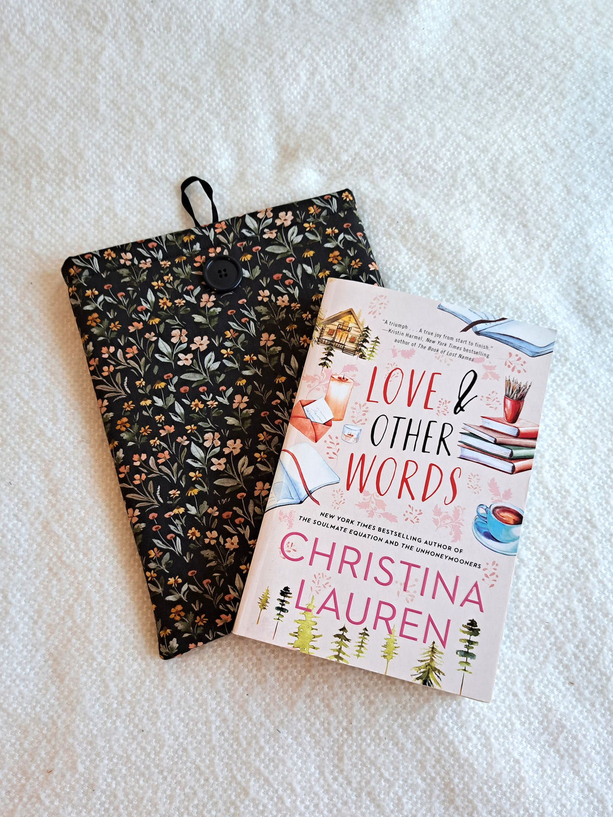 small booksleeve layed out on a white blanket. The print has a dark background with dense small flowers and leaves in a non structured pattern. The flower colors consist of sage, light pink, yellow, terracotta, and red. Black button on front with black elastic on the back. The book Love & Other Words by Christina Lauren is layed on top of the booksleeve at an angle