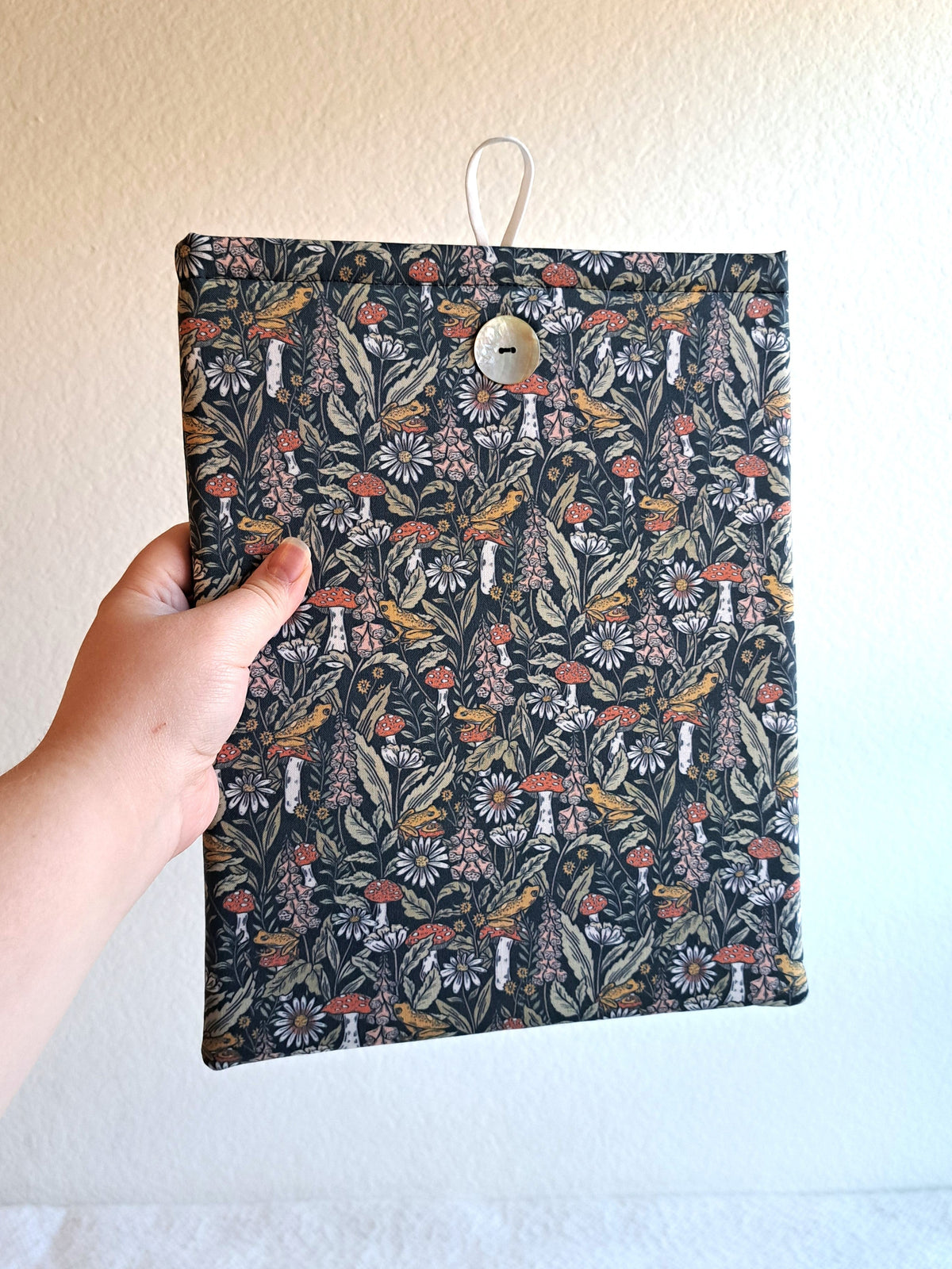 hand holding the booksleeve out. print is a dense visual print with mushrooms, leaves, and florals with small yellow frogs spread out throught the print. Colors are more pastely with blues, teals, greens, pink, and yellow. Akoya shell button on front and white elastic on back