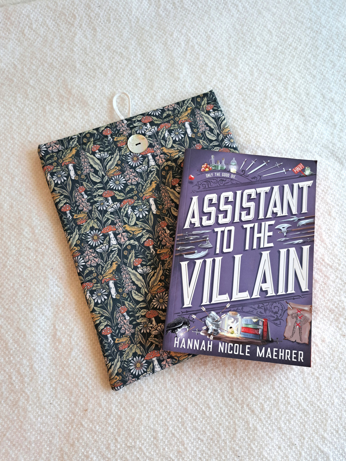 sleeve is layed out on a white blanket the print is a dense visual print with mushrooms, leaves, and florals with small yellow frogs spread out throught the print. Colors are more pastely with blues, teals, greens, pink, and yellow. Akoya shell button on front and white elastic on back. The book Assistant to the villain is layed out on top of the sleeve