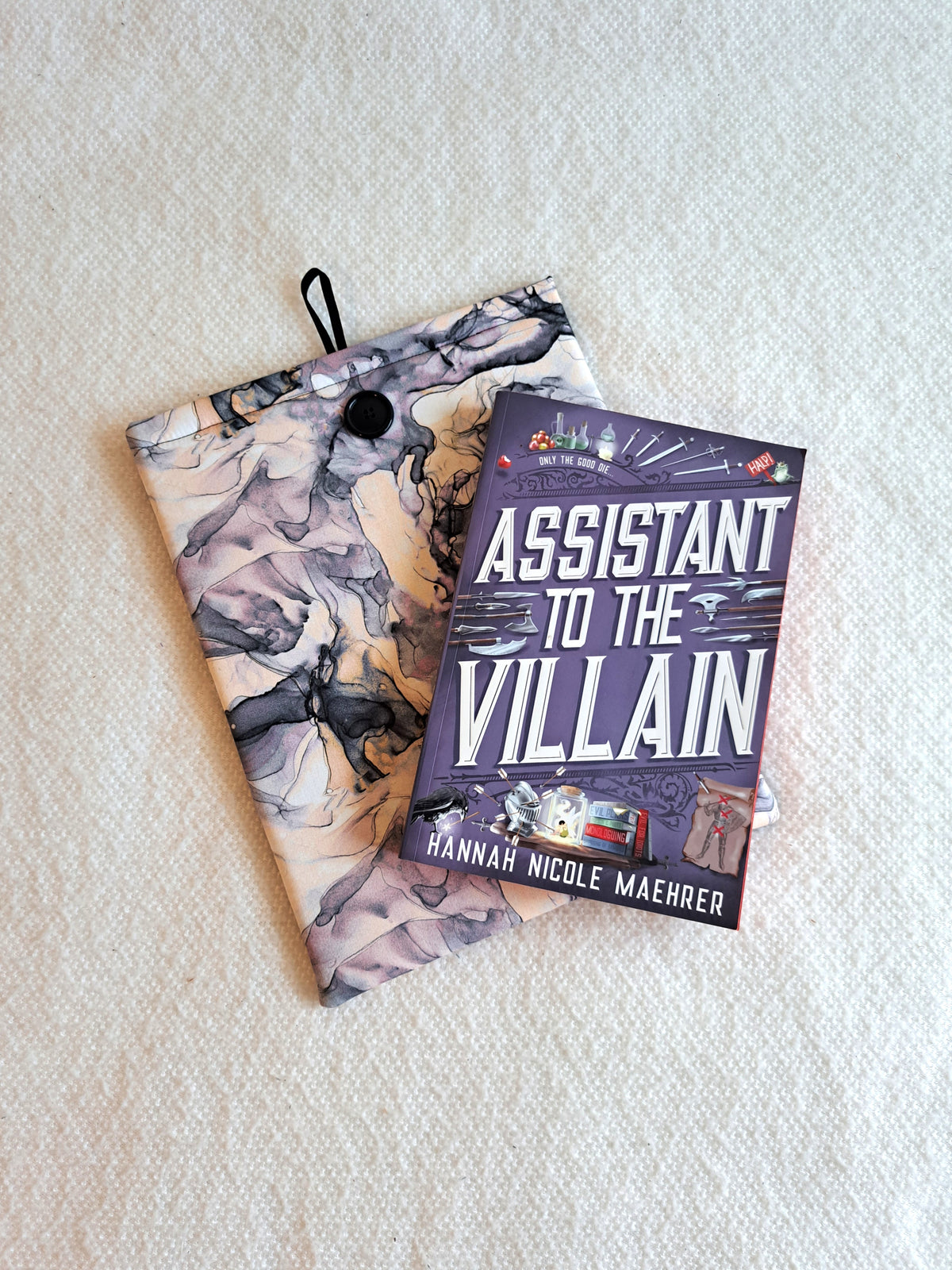 sleeve is layed out on a white blanket. The print is fluid watercolor in various purples, cream, white, and black. Purple is the leading color. Black shell button on front and black elastic on back. The book Assistant to the Villain is layed on top of the sleeve at an angle