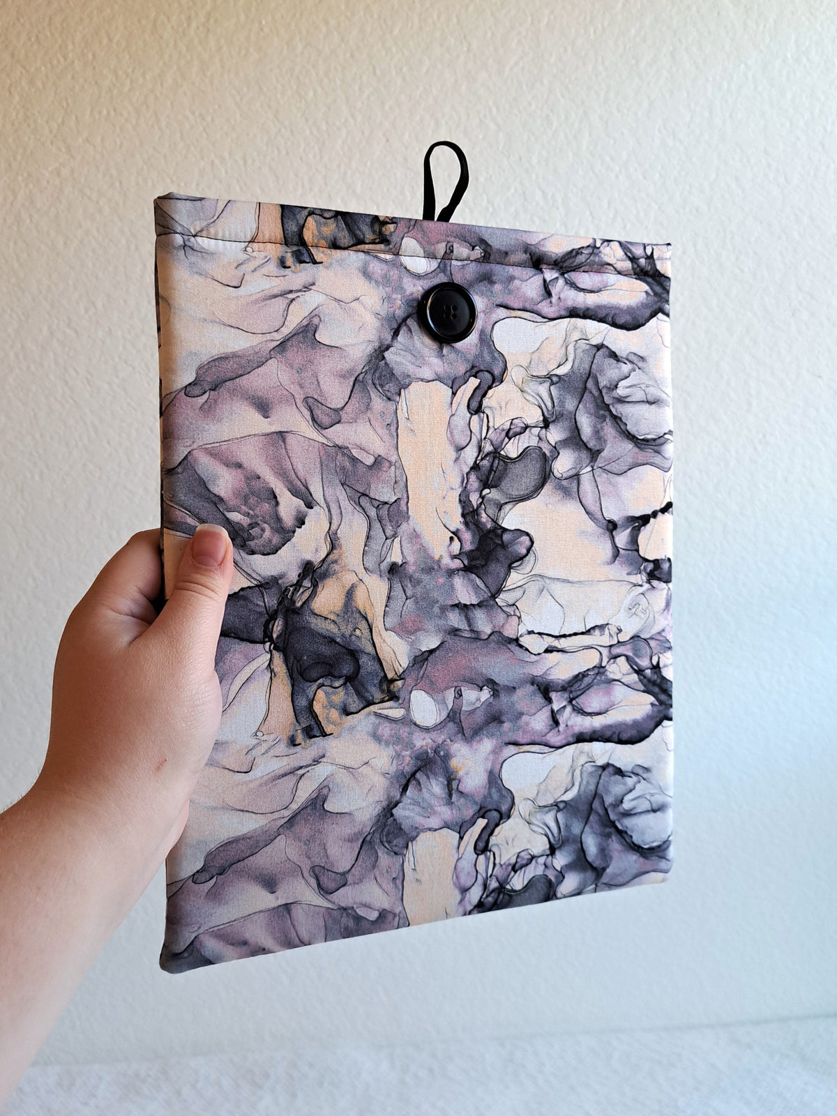 hand holding the booksleeve out. print is fluid watercolor in various purples, cream, white, and black. Purple is the leading color. Black shell button on front and black elastic on back