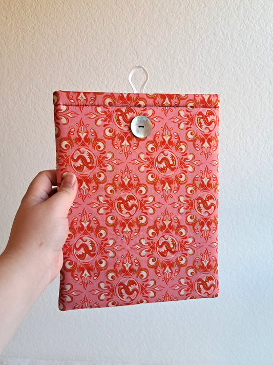 Hand holding out the booksleeve. Print is of decorative dragons and swirls in rosy and pink colors. Button is shiny Akoya shell. White elastic loop on the back