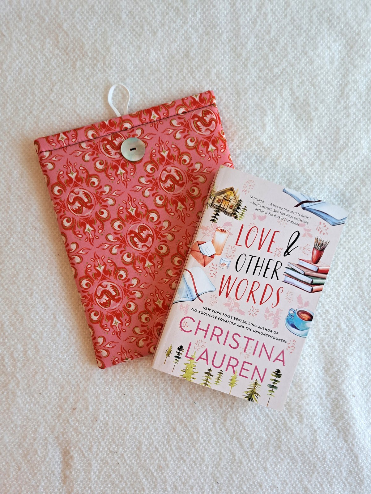 booksleeve is layed out on a white blanket. Print is of decorative dragons and swirls in rosy and pink colors. Button is shiny Akoya shell. White elastic loop on the back. The book Love & Other Words is layed out on top at an angle