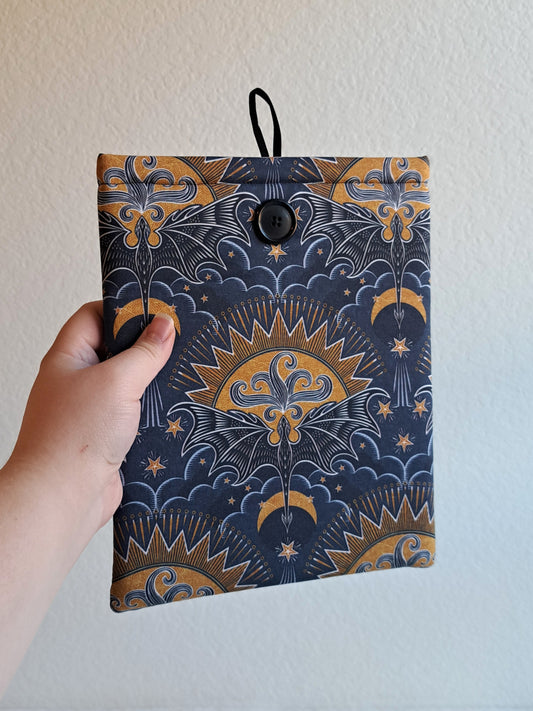 hand holding the booksleeve out. print is a repeated motiff of a blue dragon with its wings spread out with a geometric sun in gold yellow colors over it, illustrative clouds under it. and various stars and lines around it giving the look of the dragon flying.black button on front and black elastic on back