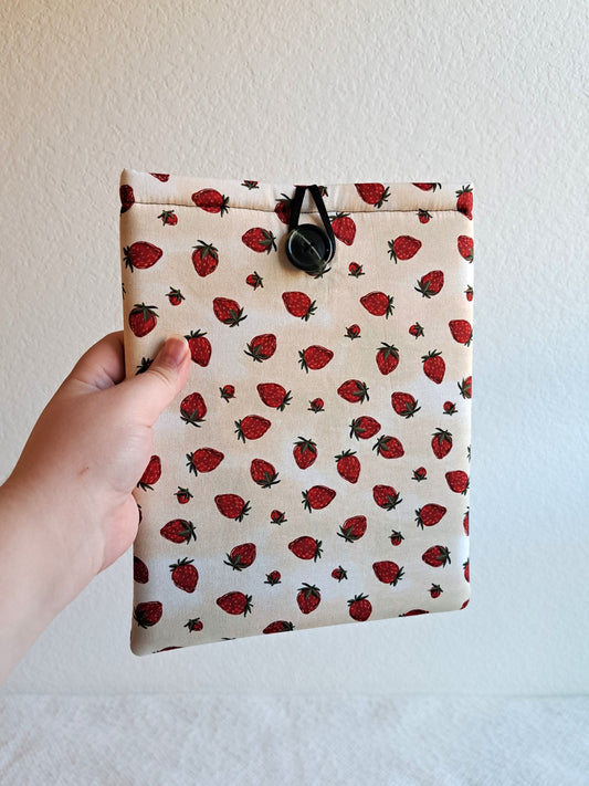 hand holding the booksleeve out. print is a light cream background with strawberries scattered across the fabris. Black button on front and black elastic on back