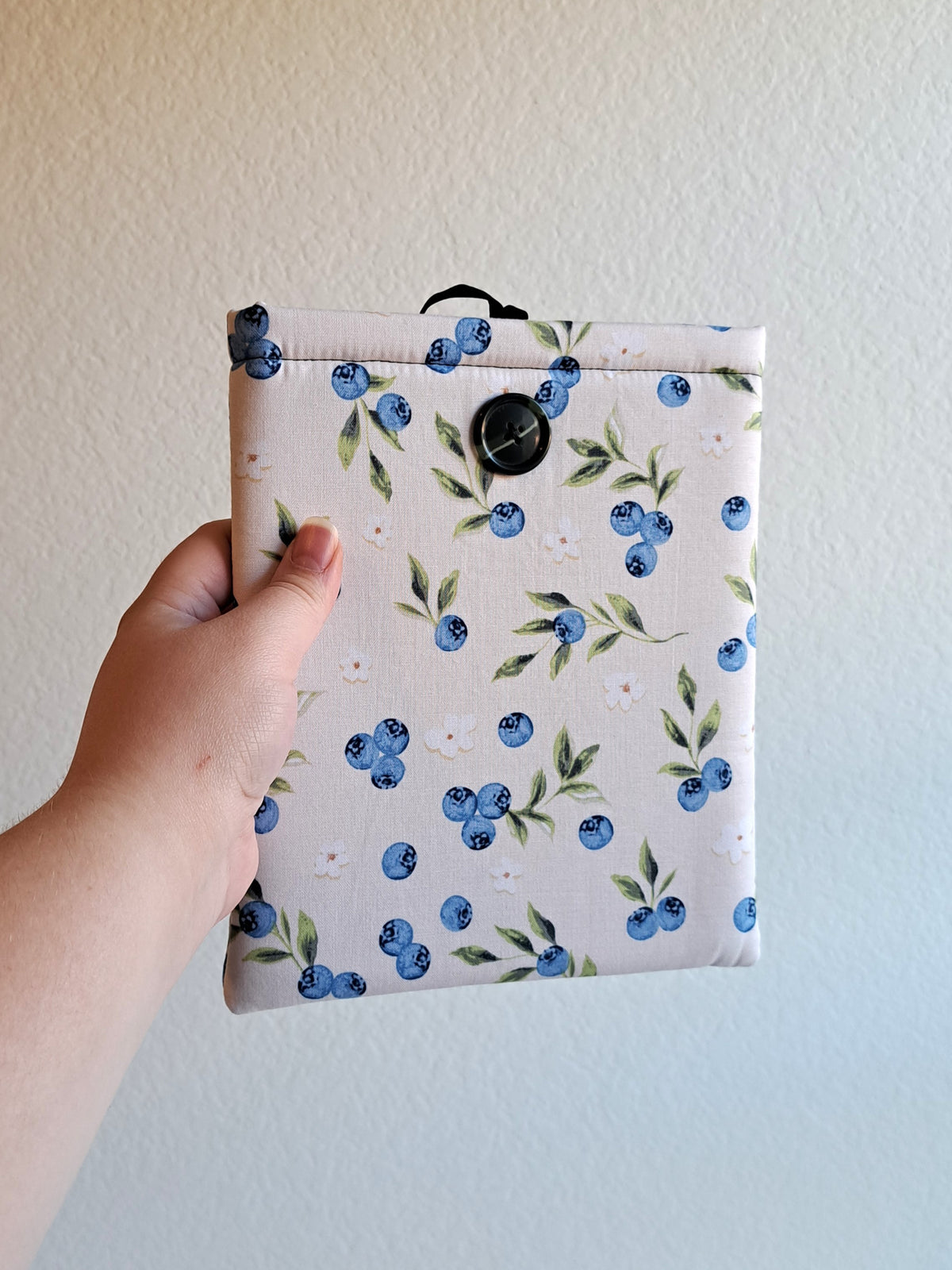 a hand is holding the kindle sleeve out. print is a light cream background with blueberries, leaves, and small white flowers spread out around the sleeve. Black button on front and black elastic on back