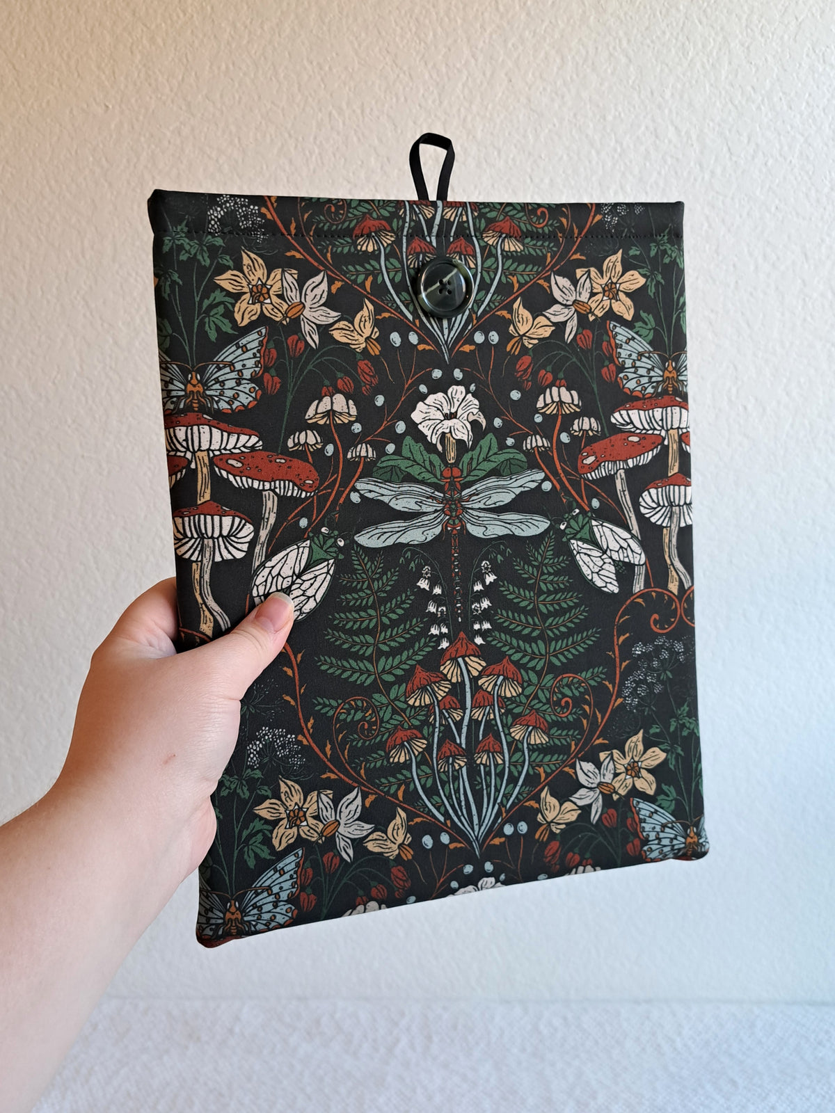 hand holding the booksleeve out. print is on a dark background, a dragonfly motiff in the center surrounded by various mushrooms and plants in a dark earthy color palette. Black button on front and black elastic on back