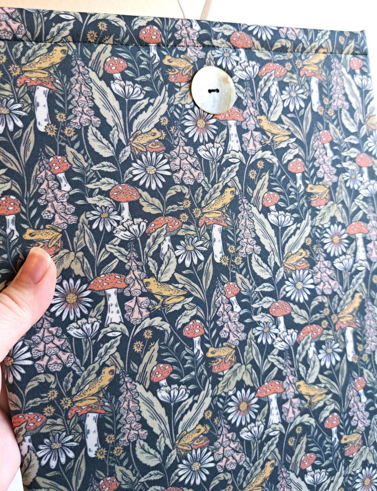 close up of a hand holding the booksleeve out. print is a dense visual print with mushrooms, leaves, and florals with small yellow frogs spread out throught the print. Colors are more pastely with blues, teals, greens, pink, and yellow. Akoya shell button on front and white elastic on back