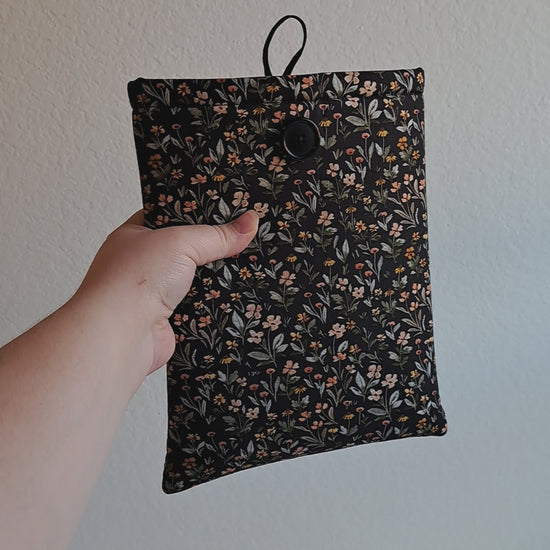 video of the small booksleeve held out by a hand. The print has a dark background with dense small flowers and leaves in a non structured pattern. The flower colors consist of sage, light pink, yellow, terracotta, and red. Black button on front with black elastic on the back