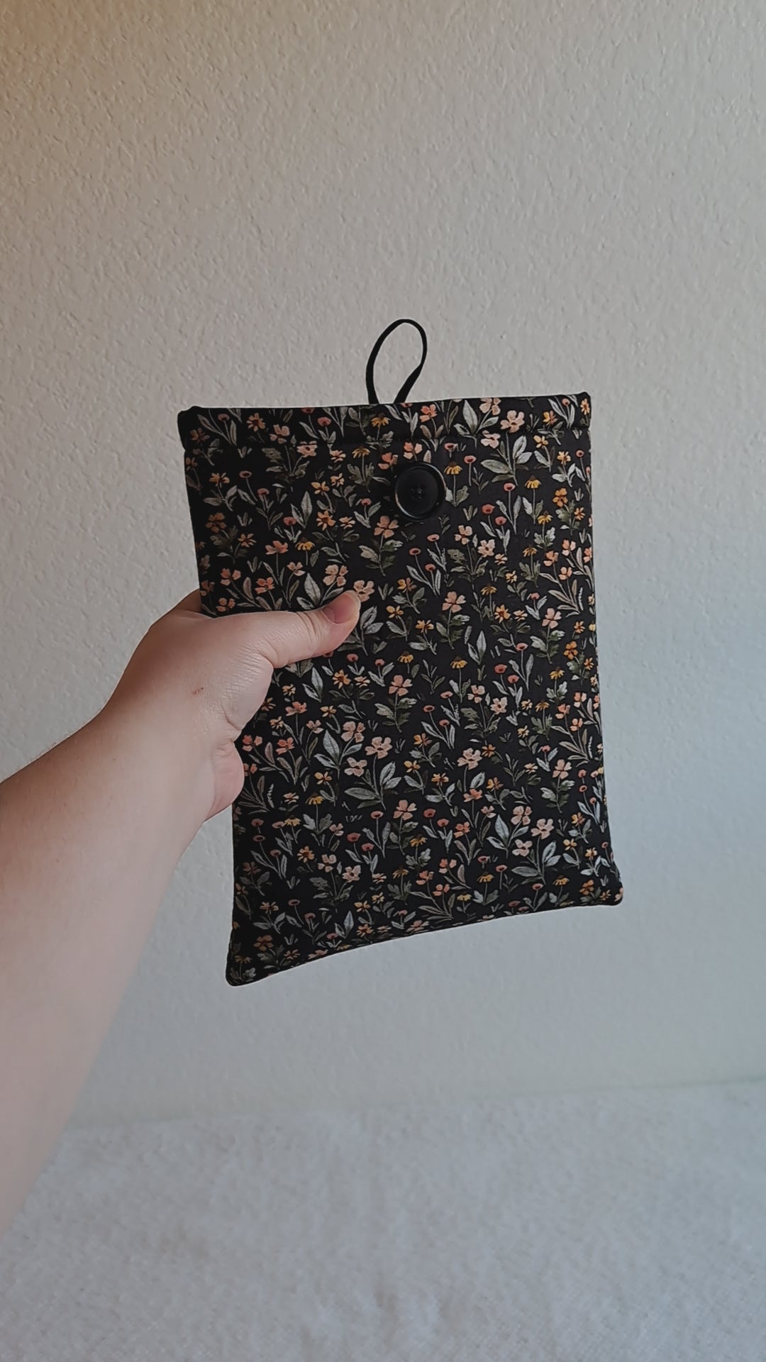 video of the small booksleeve held out by a hand. The print has a dark background with dense small flowers and leaves in a non structured pattern. The flower colors consist of sage, light pink, yellow, terracotta, and red. Black button on front with black elastic on the back