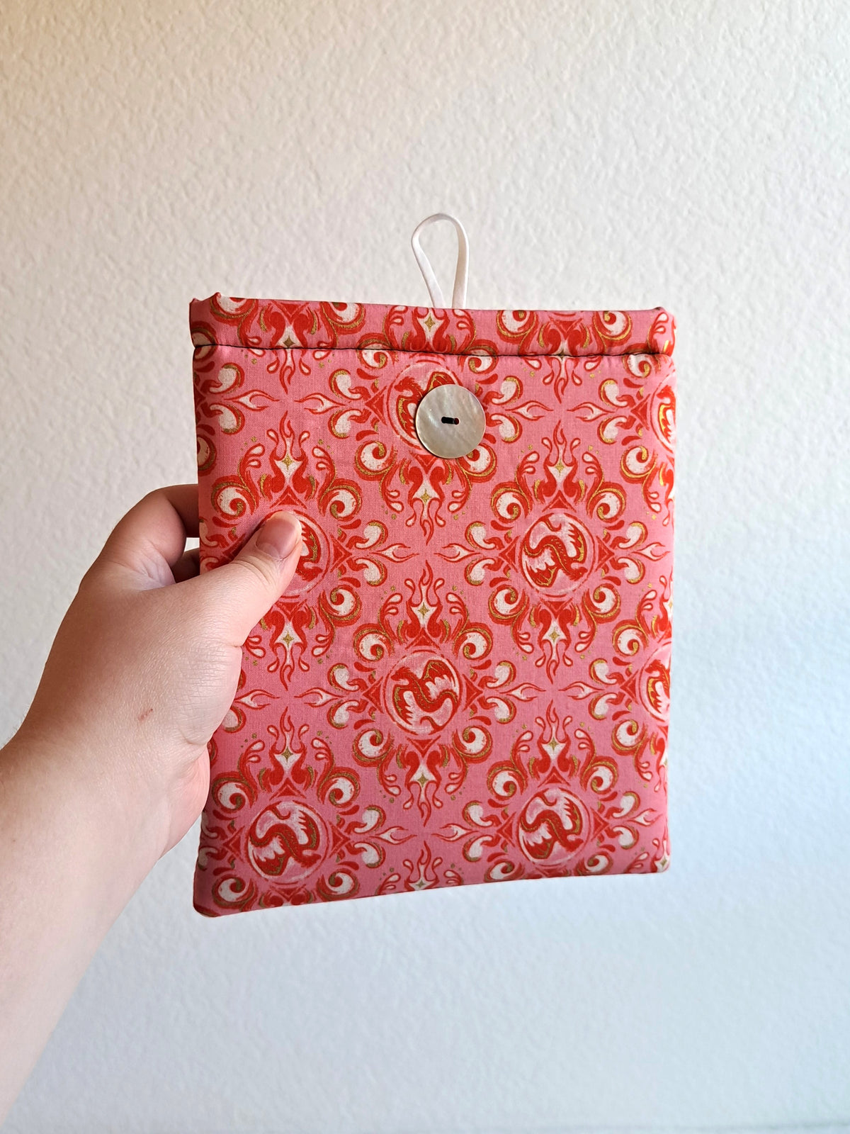 Hand holding out the kindle sleeve. Print is of decorative dragons and swirls in rosy and pink colors. Button is shiny Akoya shell. White elastic loop on the back