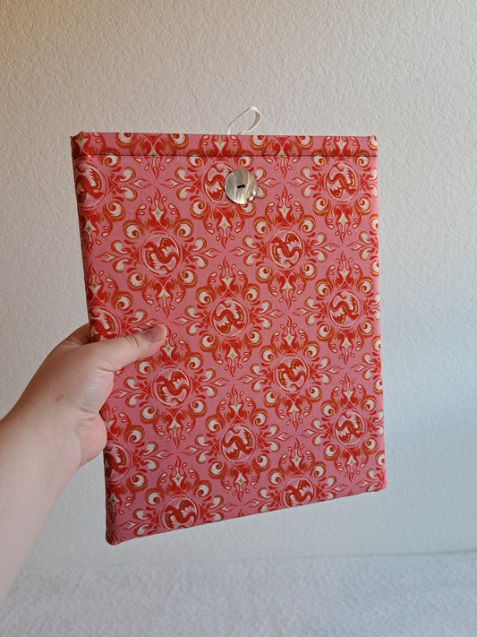 Hand holding out the booksleeve. Print is of decorative dragons and swirls in rosy and pink colors. Button is shiny Akoya shell. White elastic loop on the back