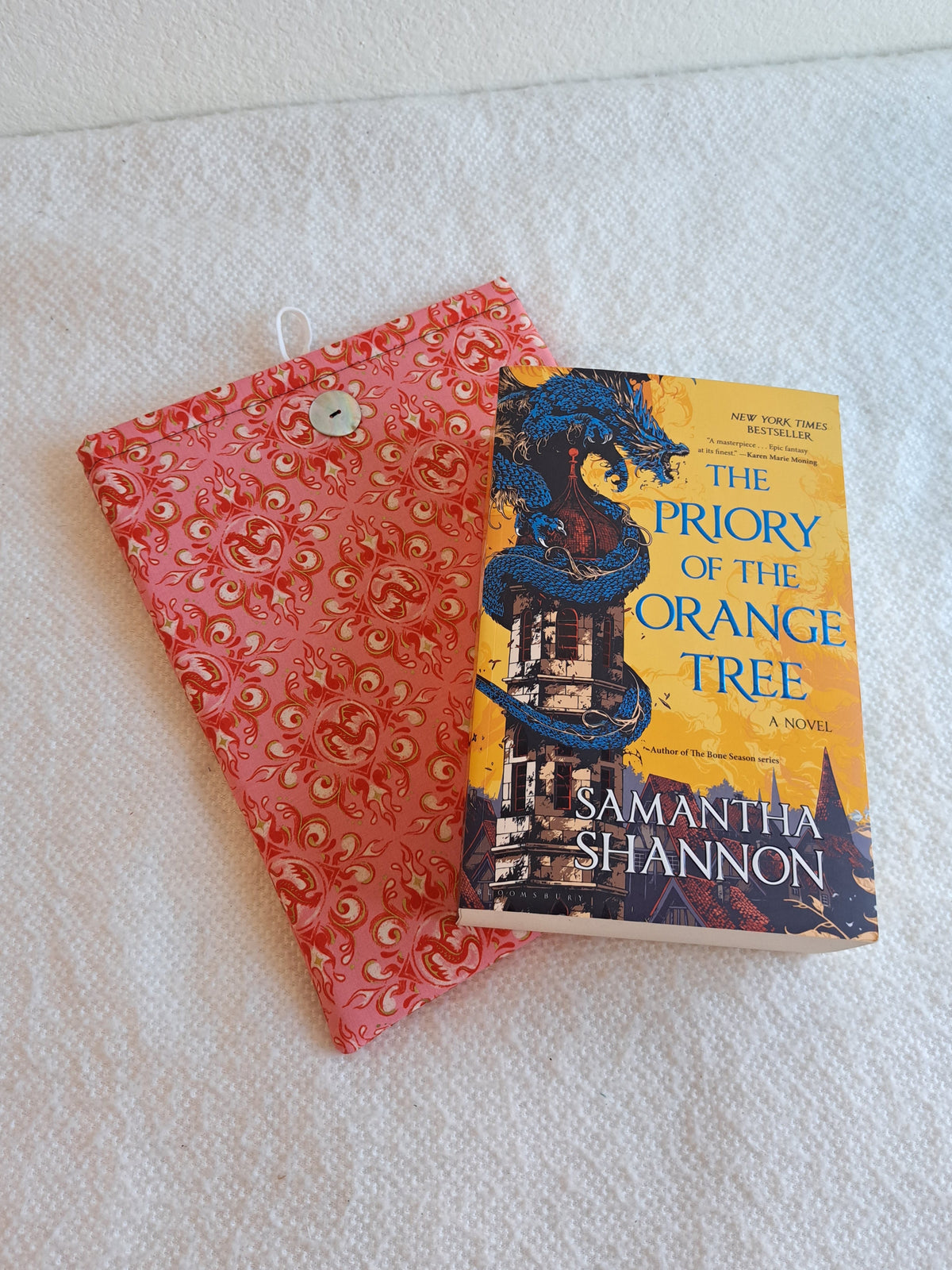 Booksleeve place on white blanket. Print is of decorative dragons and swirls in rosy and pink colors. Button is shiny Akoya shell. White elastic loop on the back. The book Priory of the orange tree is set on top