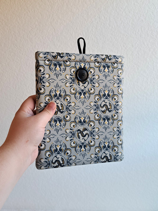 hand holding the kindle sleeve out. print is a light gray background with navy, black and gold metallic illustrative graphics of dragons and swirl motiffs. Black button on front and black elastic on back
