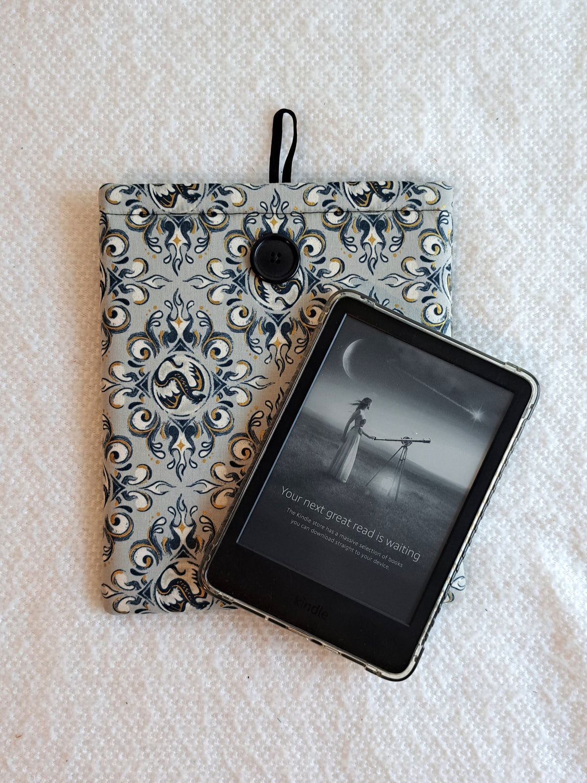 the kindle sleeve is laying on a white blanket. the print is a light gray background with navy, black and gold metallic illustrative graphics of dragons and swirl motiffs. Black button on front and black elastic on back
A kindle is layed on top of the sleeve at an angle