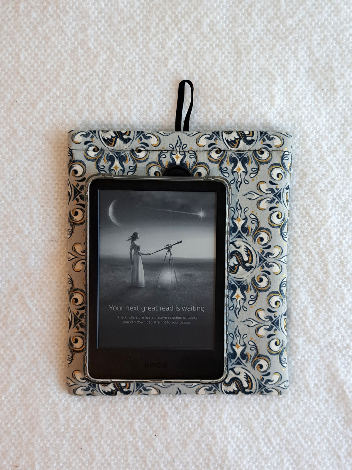 the kindle sleeve is laying on a white blanket. the print is a light gray background with navy, black and gold metallic illustrative graphics of dragons and swirl motiffs. Black button on front and black elastic on back
A kindle is layed on top of the sleeve