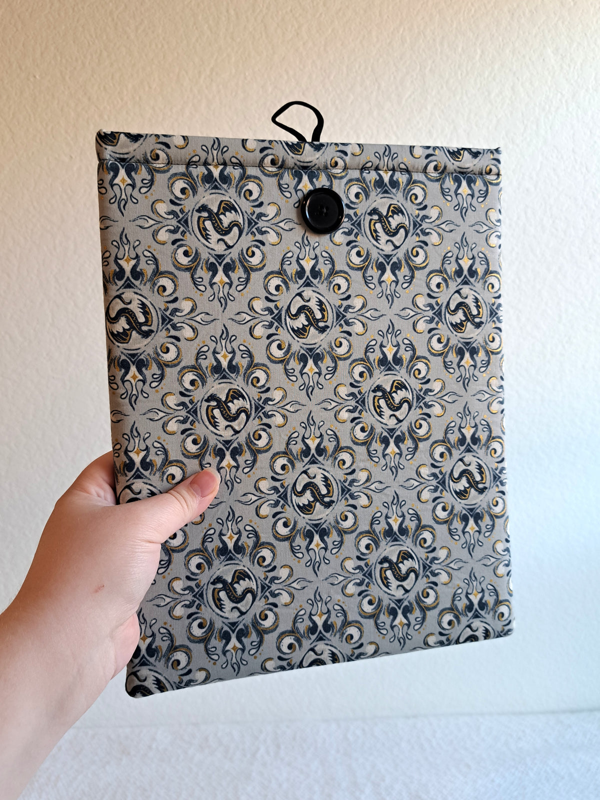 hand holding the booksleeve out. print is a light gray background with black and gold metallic illustrative graphics of dragons and swirl motiffs. Black button on front and black elastic on back