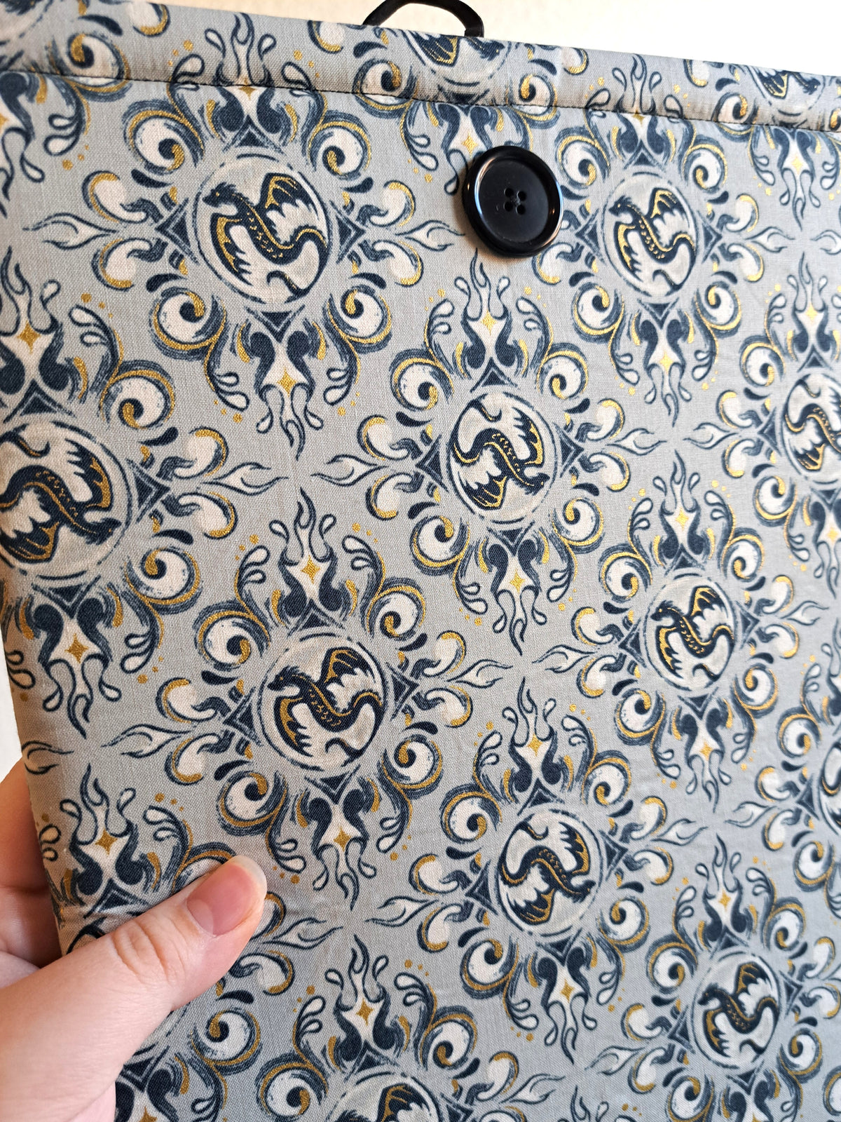 close up of hand holding the booksleeve out. print is a light gray background with black and gold metallic illustrative graphics of dragons and swirl motiffs. Black button on front and black elastic on back