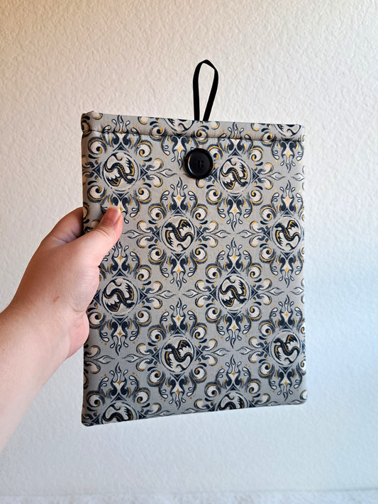 hand holding the booksleeve out. print is a light gray background with black and gold metallic illustrative graphics of dragons and swirl motiffs. Black button on front and black elastic on back