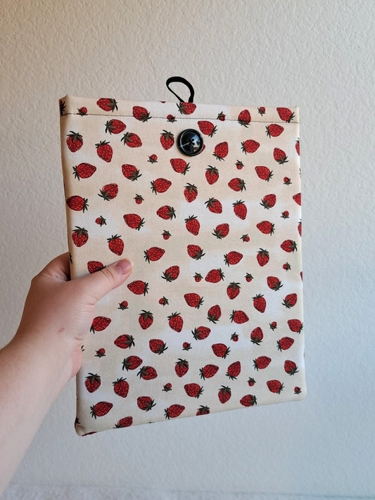 hand holding the booksleeve out. print is a light cream background with strawberries scattered across the fabris. Black button on front and black elastic on back