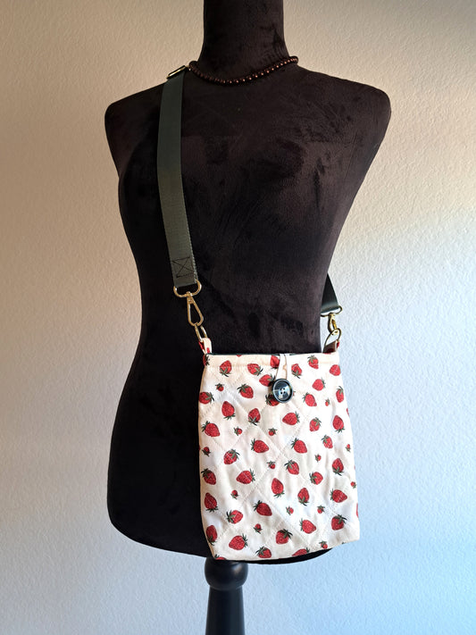 Bag is hanging on a black dress form with a white background. the print of the bag  is a light cream background with strawberries scattered across the fabric. green button on front and black elastic on back. Gold hardware and a sage green strap.