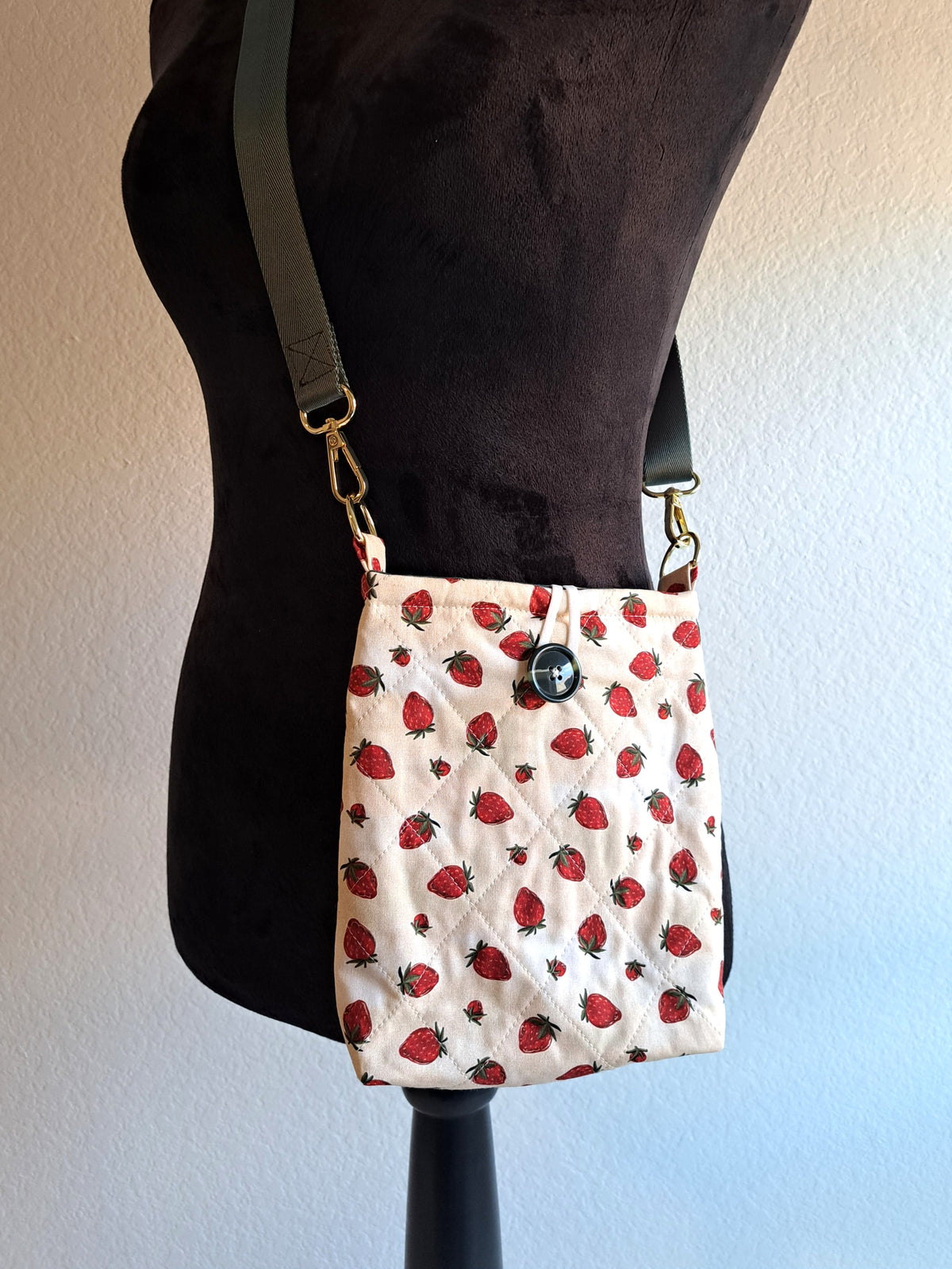 the bag is hanging on a black dressform with a white background. the print of the bag is a light cream background with strawberries scattered across the fabric. It is quilted in a diamond pattern. green button on front and black elastic on back. Gold hardware and a sage green strap