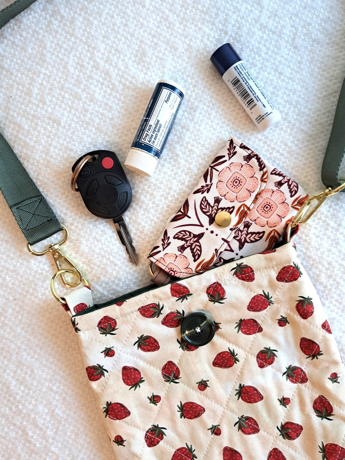 the bag is layed out on a white blanket with keys, chapstick and a wallet coming out of it. the print of the bag is a light cream background with strawberries scattered across the fabric. It is quilted in a diamond pattern. green button on front and black elastic on back. Gold hardware and a sage green strap