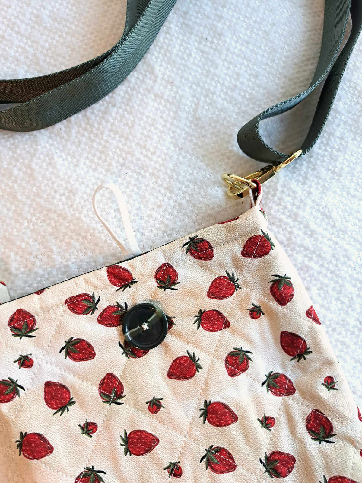 the bag is layed out on a white blanket. the print of the bag is a light cream background with strawberries scattered across the fabric. It is quilted in a diamond pattern. green button on front and black elastic on back. Gold hardware and a sage green strap